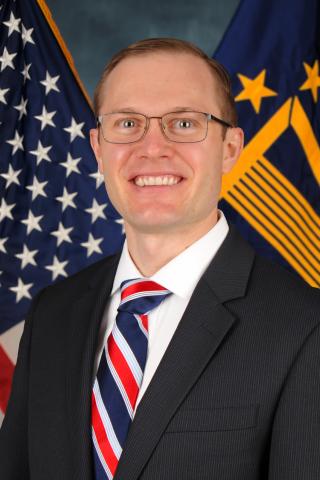 Man in a suit and red, white, and blue tie in front of American and DHS flags.