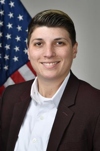 A smiling woman in a white shirt and brown blazer stands in front of an American flag.