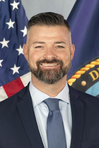 Man in a navy suit and blue tie smiling, with American and Department of Defense flags behind him.