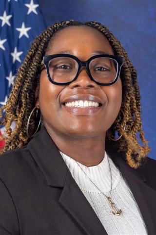 Woman with braids wearing glasses and a black blazer, smiling in front of a US flag.