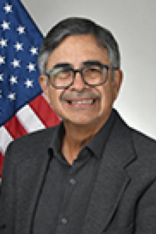 Portrait of man in black jacket and glasses smiling in front of an American flag.