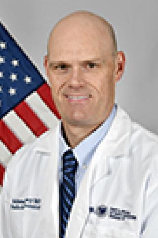 A bald man in a white lab coat and blue tie stands in front of an American flag.