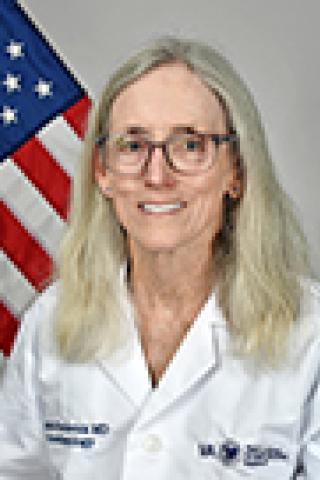 Woman in white lab coat smiling, with American flag in background.