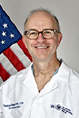 Smiling man in a lab coat with an American flag behind him.