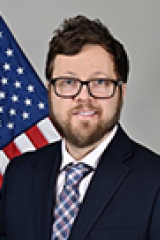 Man in a suit and tie smiling, with an American flag in the background.