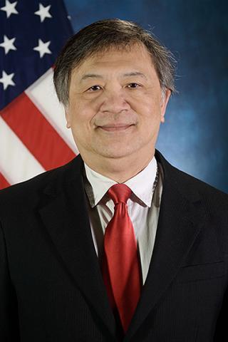 A man in a black suit and red tie stands in front of an American flag.