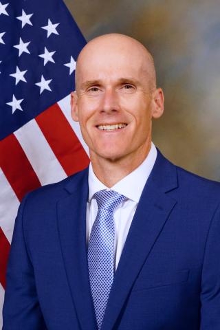 Smiling man in blue suit and tie standing in front of American flag.