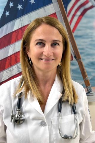Woman with blonde hair in white lab coat with stethoscope around her neck in front of an American flag.