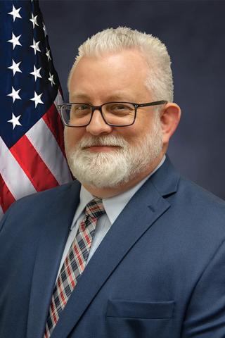 Man with white hair and beard, wearing a blue suit and red tie, standing by an American flag.