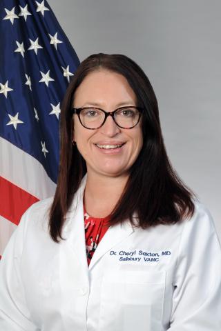 Dr. Cheryl Sexton smiling in a white lab coat with the American flag behind her.