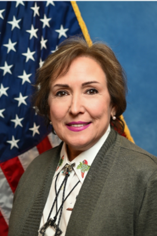 Woman in gray jacket and white shirt smiling, with American flag in background.