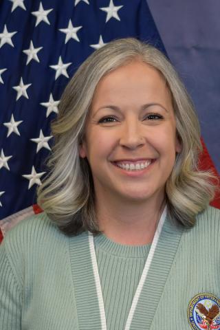 Woman with gray hair smiling in front of US flag, wearing green shirt and white lanyard.