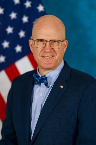 Man in suit and bow tie smiling in front of American flag.