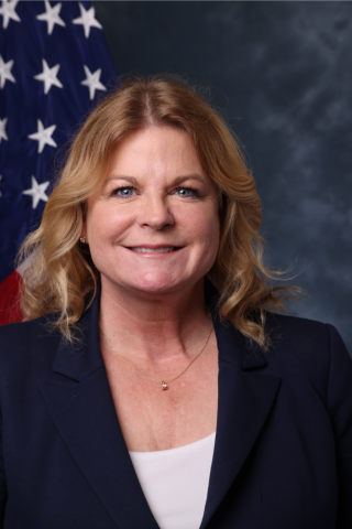Portrait of a woman in a navy blazer and white shirt, smiling, with an American flag behind her.