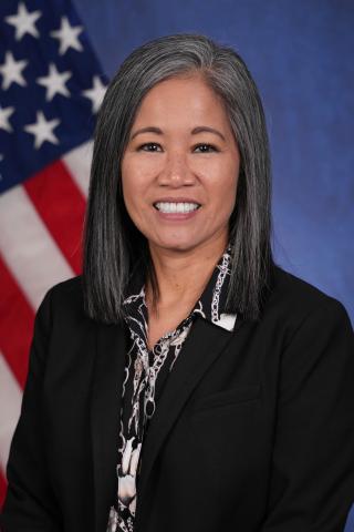 A smiling woman with gray hair, wearing a black blazer, stands in front of the American flag.