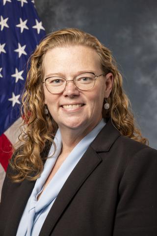 Woman with curly hair wearing glasses and a black blazer, smiling in front of an American flag.