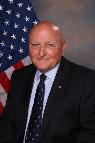 Bald man in black suit and tie standing in front of an American flag.