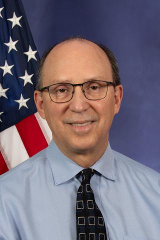 Man in blue shirt and black tie standing in front of an American flag.