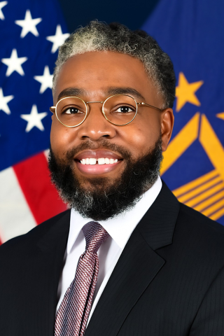 Smiling man in business suit stands in front of US and DC flags.