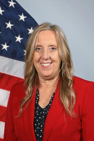 Portrait of a smiling woman in a red blazer in front of an American flag.