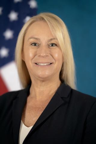 A smiling woman with blonde hair in a black blazer with an American flag in the background.