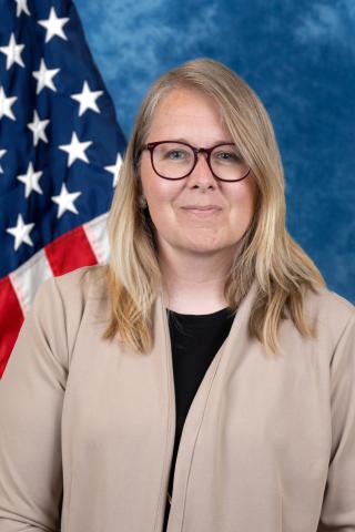 Portrait of a woman in glasses and a beige jacket standing before an American flag.