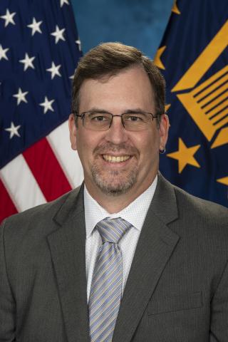 A smiling Ian Komorowski in a gray suit and striped tie stands in front of American and state flags.