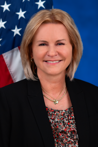 Woman with blonde hair wearing a black blazer and patterned top, smiling in front of an American flag.