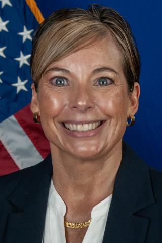 A smiling woman with short blonde hair in business attire stands in front of an American flag.