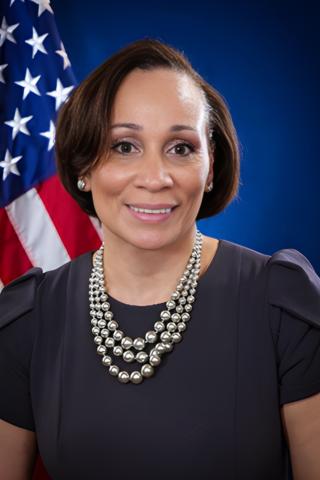A woman in a black dress and pearl necklace smiles in front of an American flag.