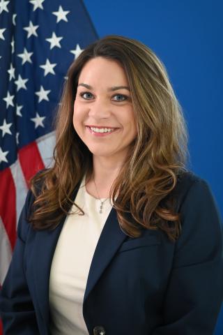 Portrait of a smiling woman in a white shirt and blue blazer, standing before a U.S. flag.