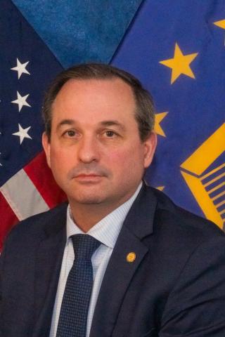 Portrait of a man in a suit with American and European Union flags in the background.