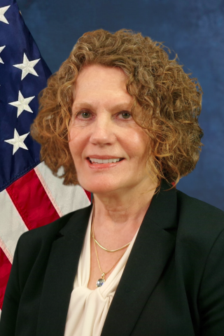 A smiling woman with curly hair in a black blazer in front of an American flag.