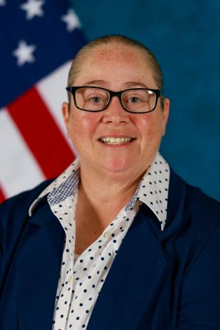 A smiling woman in glasses and a navy cardigan with an American flag behind her.