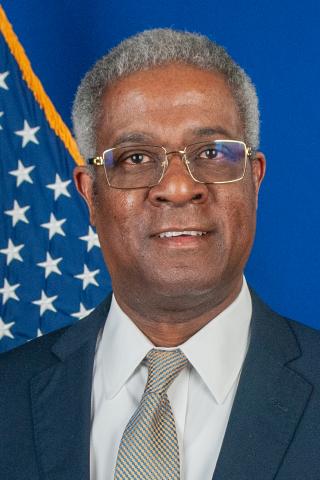 A smiling man in a suit and tie stands in front of an American flag.