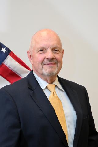 Bald man in navy suit and yellow tie standing next to an American flag.