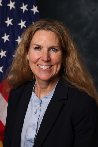 Woman with long blonde hair in a black blazer over a blue shirt standing in front of an American flag.