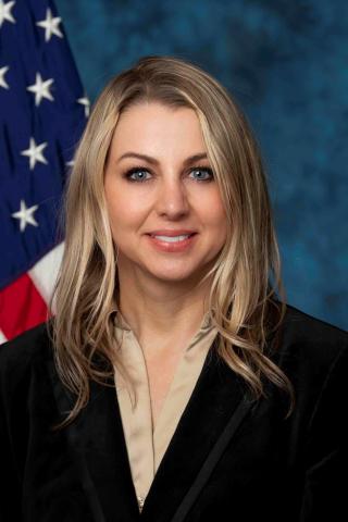 Blonde woman in black suit and white shirt smiling with American flag behind her.