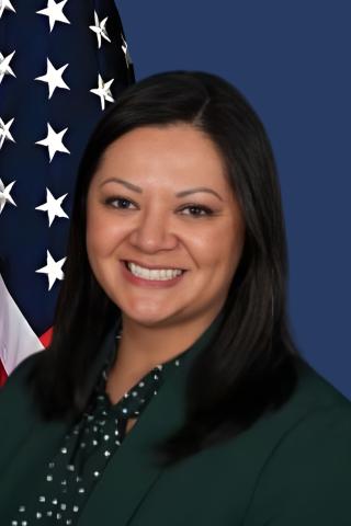 Portrait of a woman in a green blazer smiling with a US flag behind her.