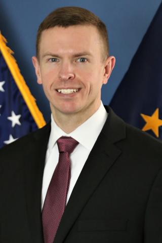Man in business attire smiling in front of USA and Department of Defense flags.