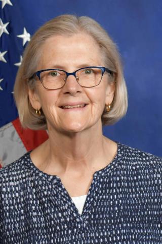 A smiling older woman wearing glasses stands in front of the US flag.