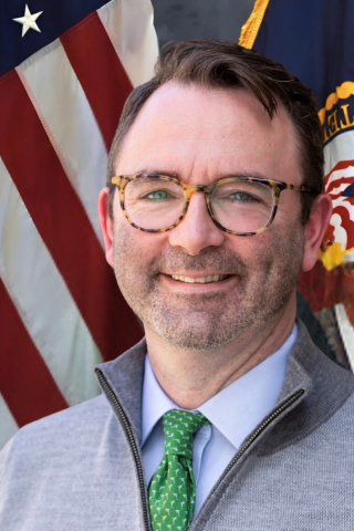 Man in gray jacket, green tie, and glasses smiling in front of American flags.