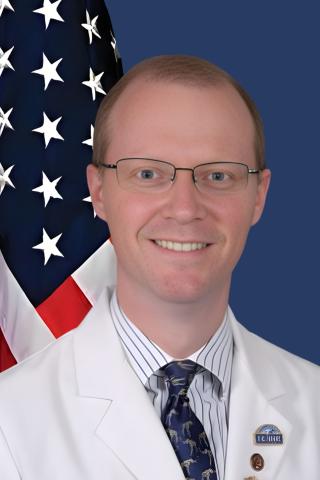 Smiling man in white lab coat and tie with American flag behind him.