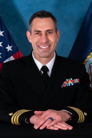 A man in navy uniform with American and military flags behind him, smiling.