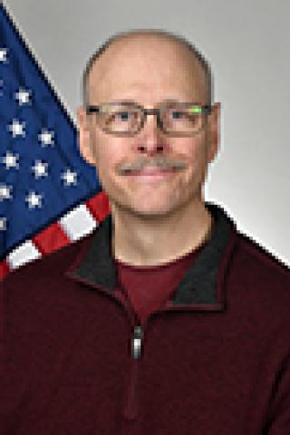 Man with mustache wearing glasses and maroon sweater in front of US flag.