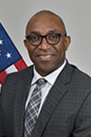 Smiling man in suit and tie standing in front of an American flag.