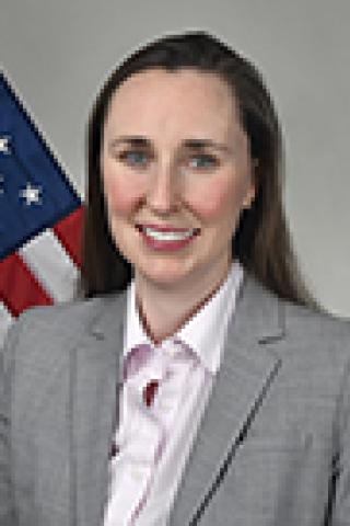 Woman in gray suit and pink shirt smiling with US flag behind her.