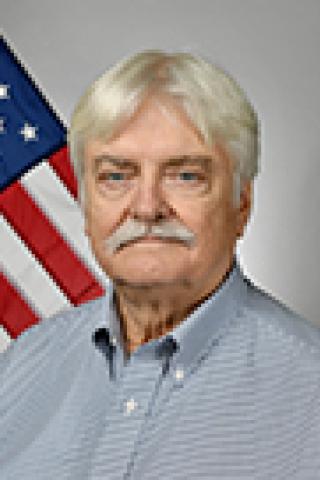 Older man with white hair and mustache, wearing blue shirt, stands in front of American flag.