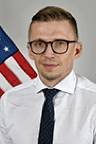 Man in a white dress shirt, black tie, and glasses stands in front of an American flag.