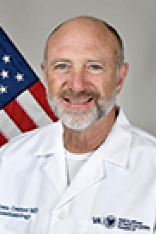 Bald man in a white lab coat smiles in front of an American flag.